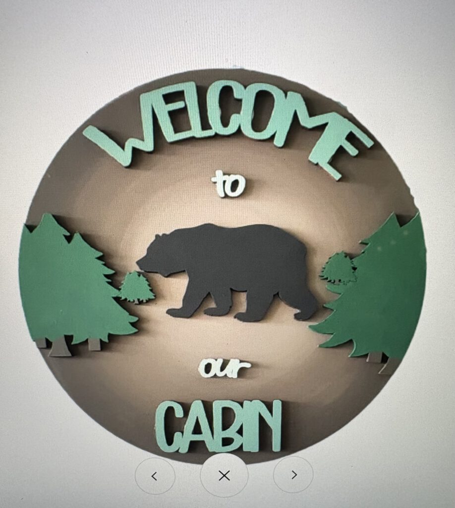 Bear Cabin - Canvas, Corks & Forks Circular badge with teal 'WELCOME TO' text above a gray bear walking among green evergreen trees on a brown circle background.