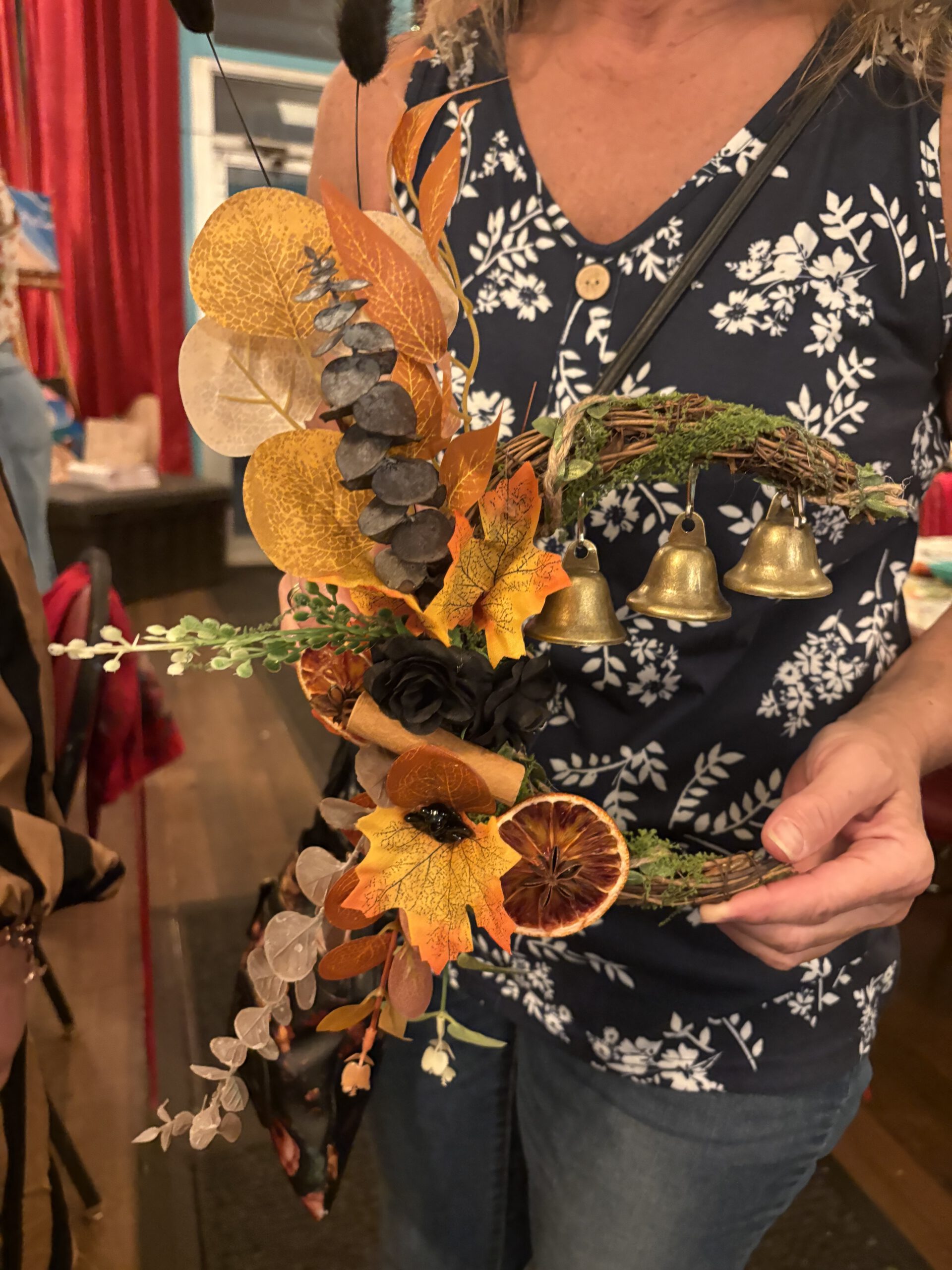 IMG_5299 - Canvas, Corks & Forks Person holding a fall-themed wreath with orange leaves, dried greenery and small brass bells indoors.